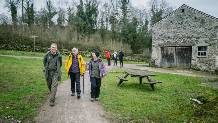 A man and two women walking the trail. To their right is an old grey stone farm building. In the background are three other walkers.